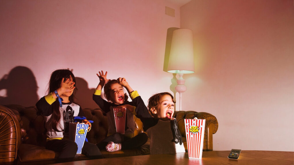 Three children sit on a brown leather couch, excitedly holding popcorn.