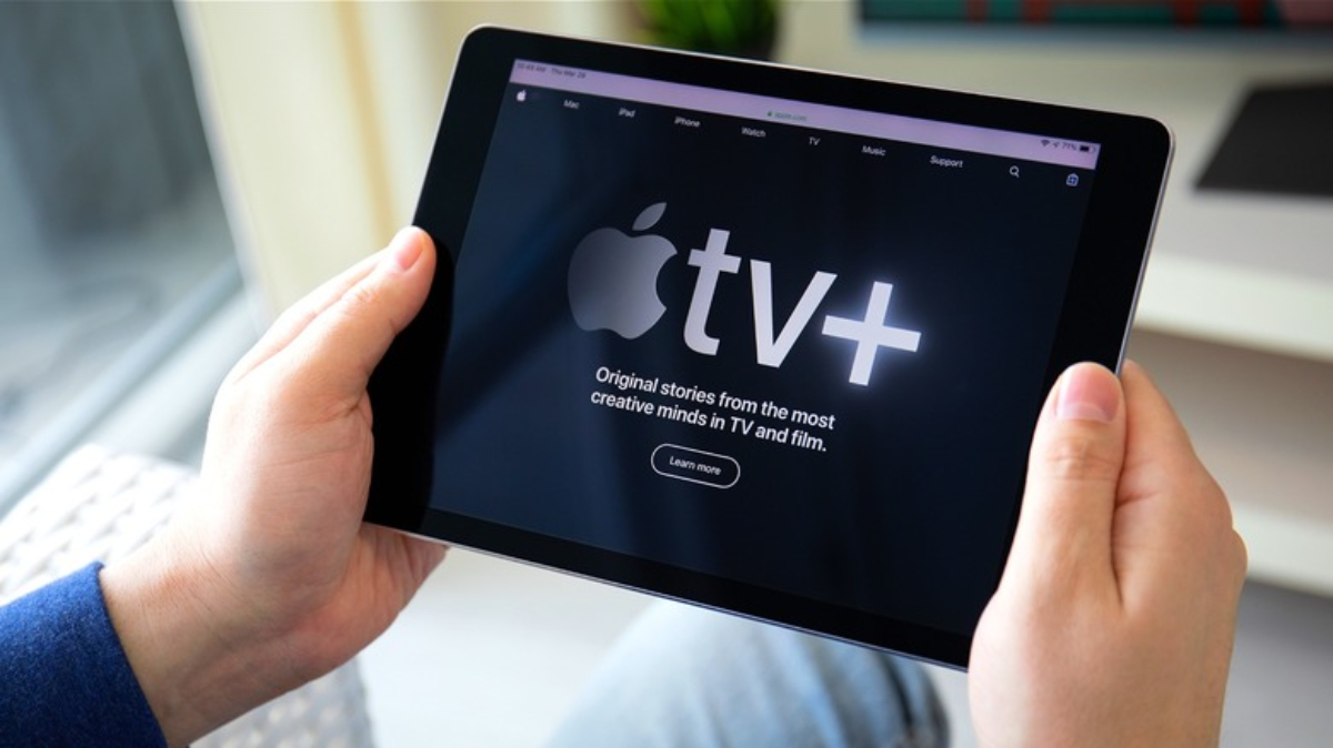 A person holds an iPad displaying the Apple TV+ homepage, featuring the slogan Original stories from the most creative minds in TV and film.