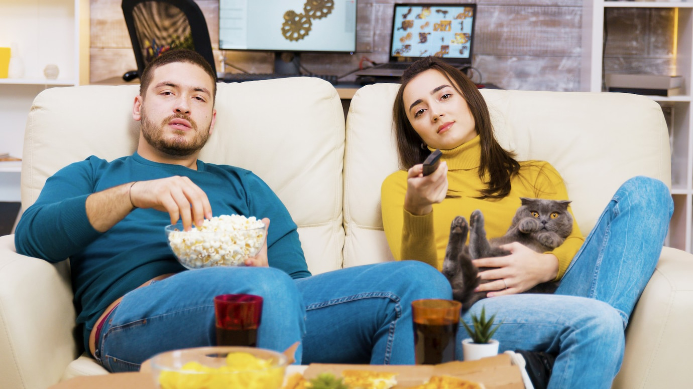 A couple sits on a couch enjoying snacks.