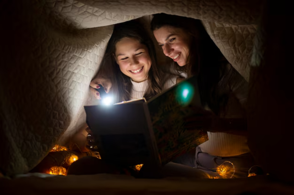 Two children reading a book with a flashlight under a blanket fort.
