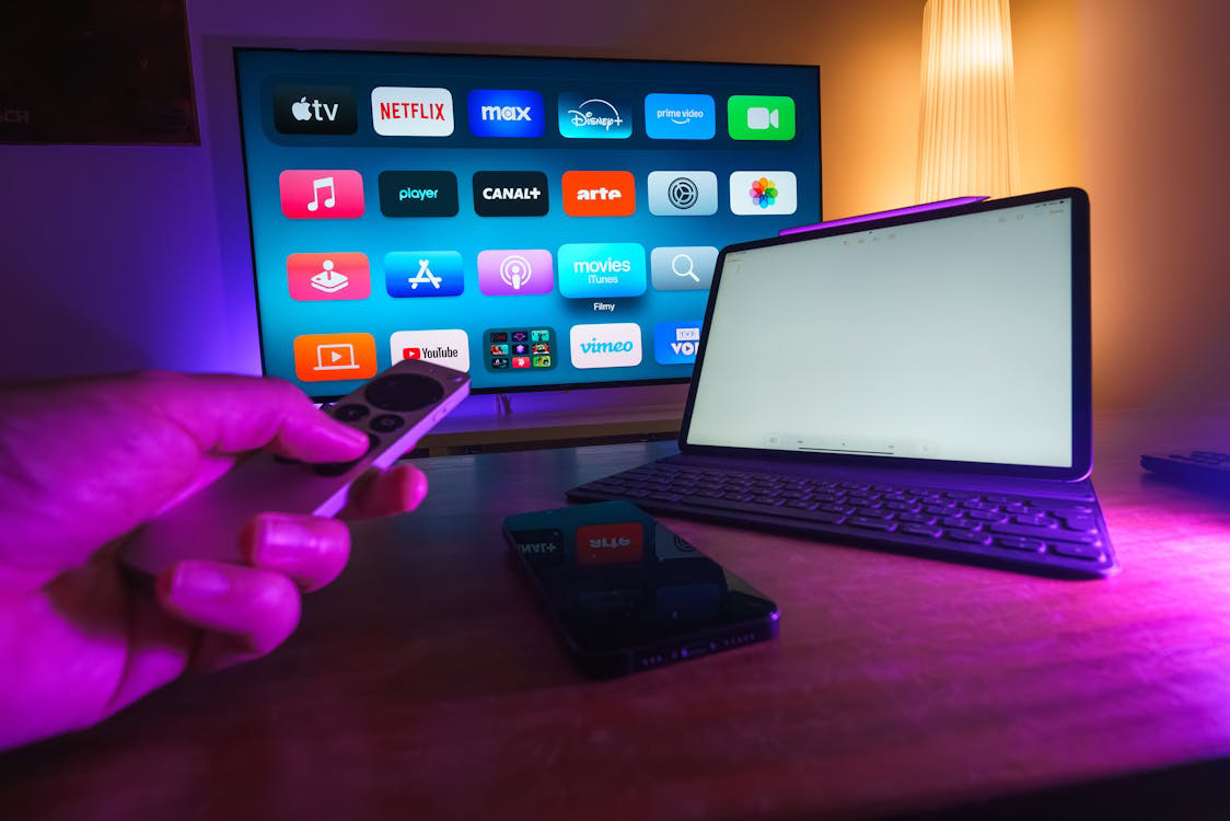 A hand holds a remote in front of a TV displaying streaming apps.