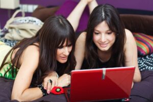 Two young women lie on a bed, intently focused on a red laptop.