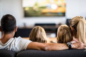 A living room scene featuring two hands holding remote controls, with a TV displaying streaming options.