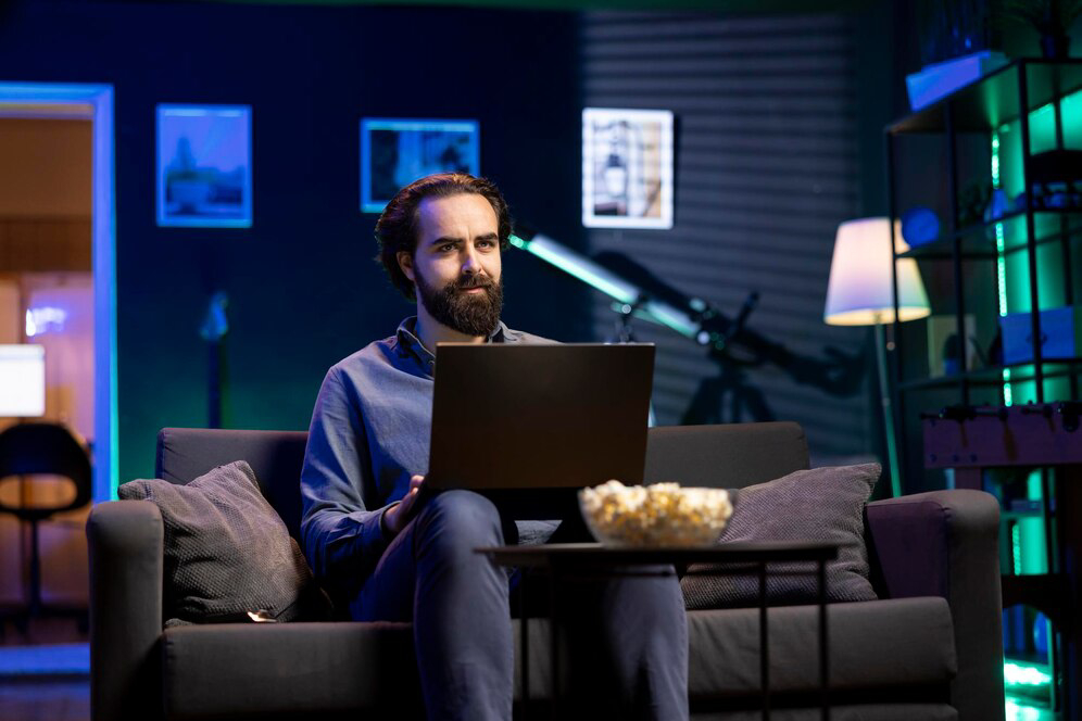 A person sits on a couch with a laptop, surrounded by cozy lighting.