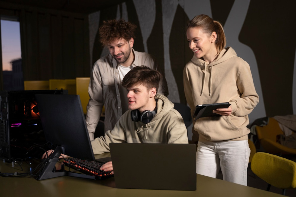 Three individuals collaborate at a desk with multiple computers.