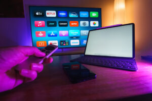 A hand holding a remote control in front of a TV displaying streaming apps.