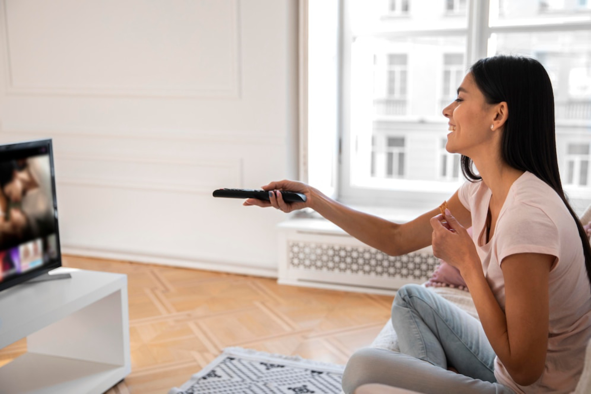 A woman sits on a couch, holding a remote and watching a screen.