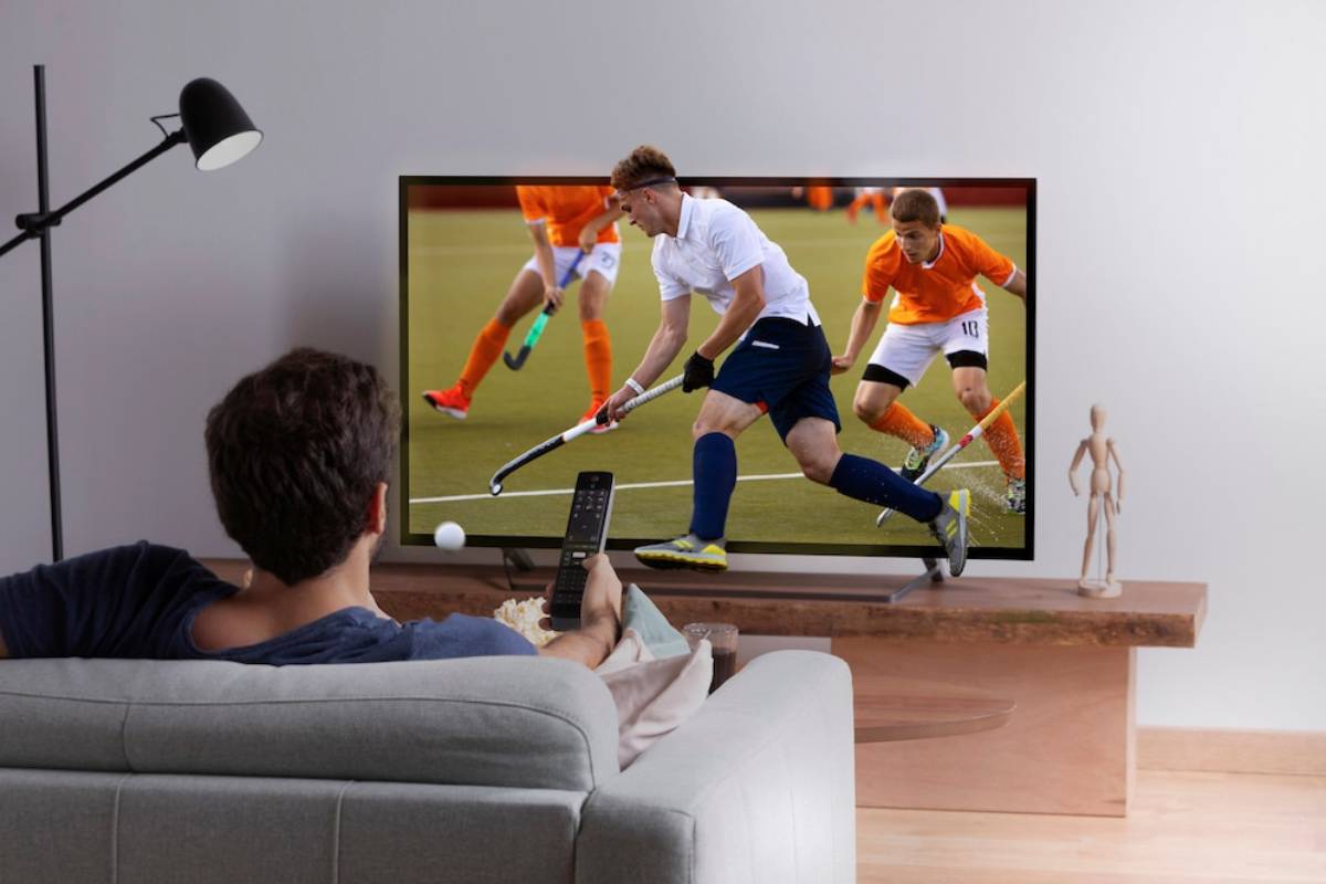 A person relaxes on a couch, watching a hockey match on a large TV with a remote in hand, enjoying snacks.