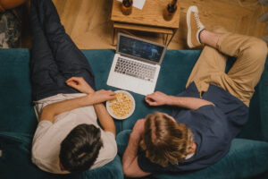 Two friends relax on a teal couch, sharing popcorn and watching a movie on a laptop.