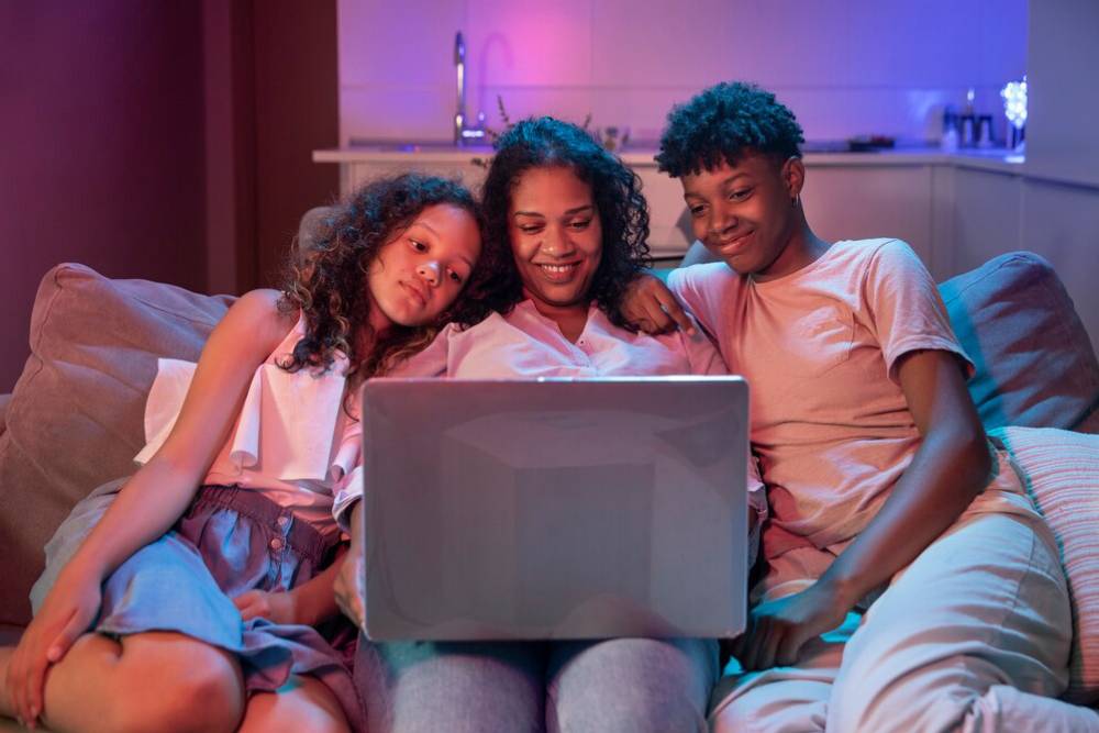 A mother and her two children relaxing on a sofa, watching a laptop together in a warmly lit room.