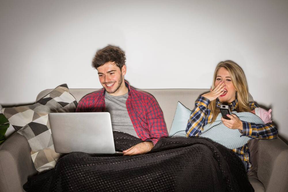 A young man and woman lounging on a sofa, laughing while using a laptop and smartphone.