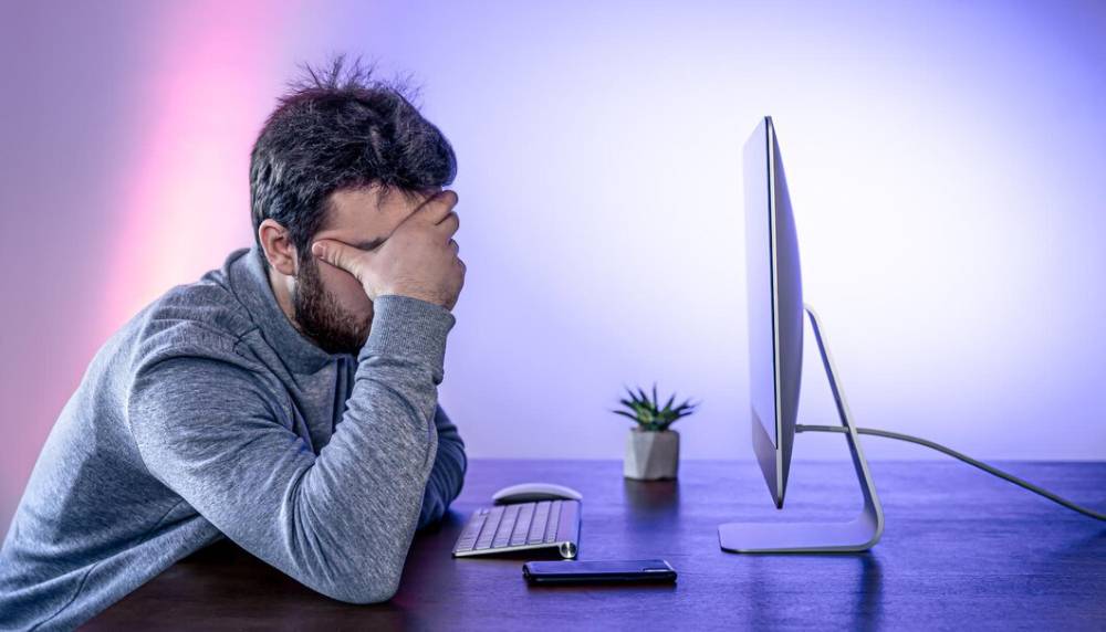 Man sitting at a desk, holding his head in frustration in front of a computer screen, with ambient purple lighting.