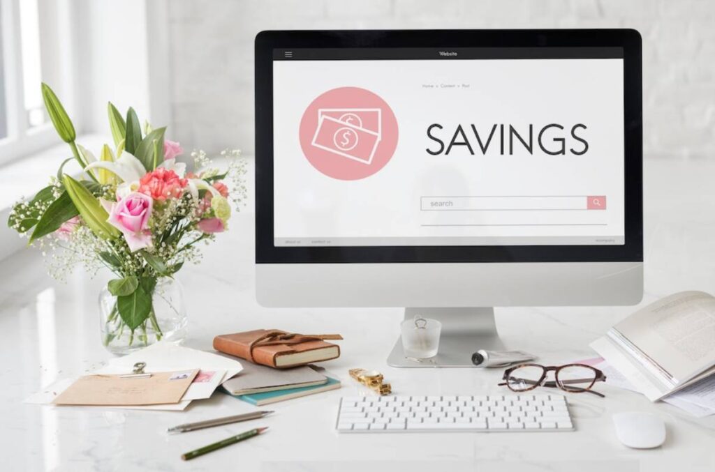 A neat workspace featuring an open computer displaying a savings webpage, surrounded by books, a bouquet of flowers, and stationery.