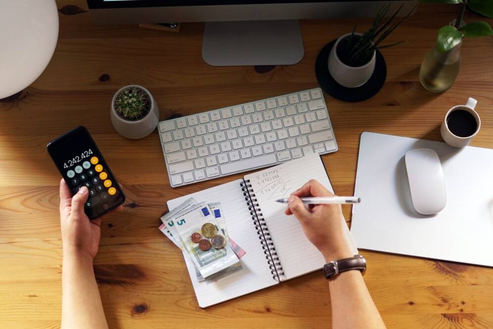A person managing finances on a calculator, with a notebook, euro cash, and keyboard on a wooden desk.