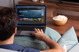 Man on a couch using a laptop displaying a shark-themed video streaming website, with a bowl of popcorn nearby.