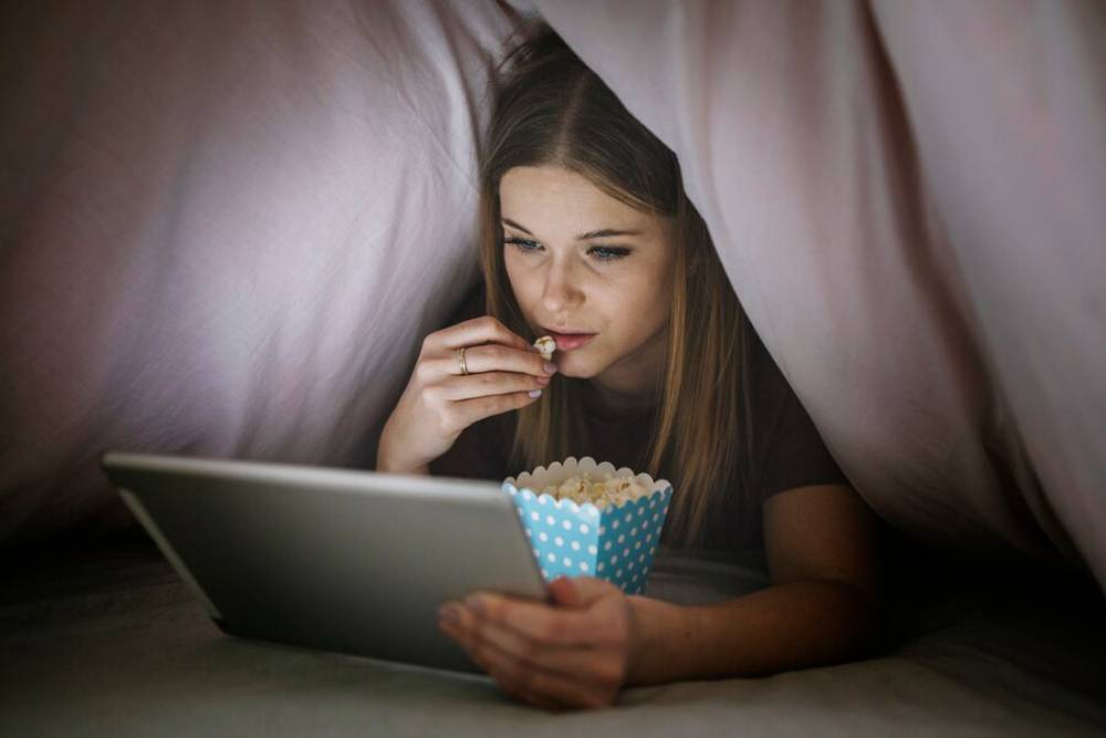Young woman eating popcorn while watching a tablet under a blanket.