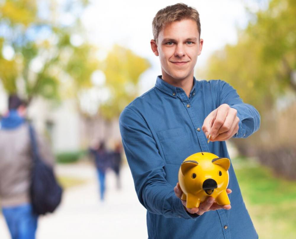 Young man in blue shirt holding a yellow piggy bank and pointing at it, outdoors with blurred people in the background.