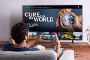 A man watches a documentary titled "Cure for the World" on TV while using his phone to stream a video, surrounded by popcorn.