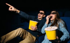 A couple watching feel good movie and looks excited, both holding large buckets of popcorn, one pointing at the screen.
