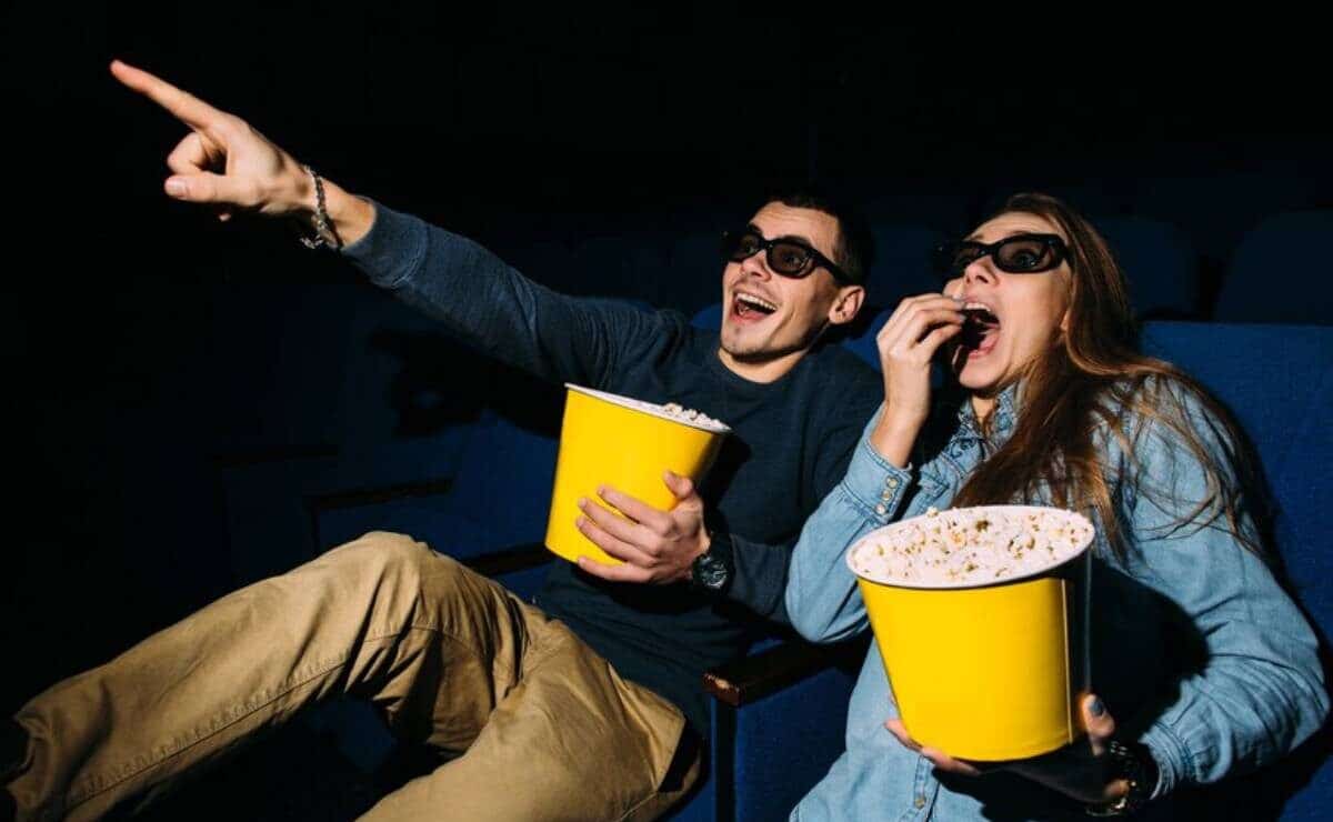 A couple watching feel good movie and looks excited, both holding large buckets of popcorn, one pointing at the screen.