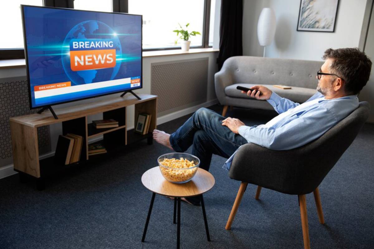 A person sits in a cozy living room, watching live breaking news on a large TV, with popcorn.