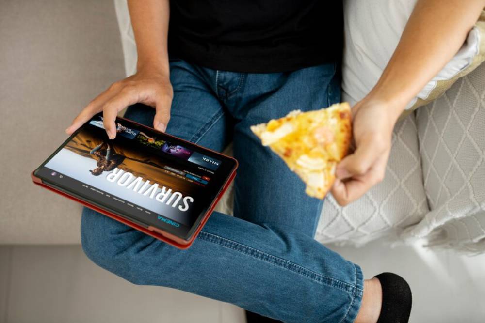 A person sitting on a couch, watching a tablet featuring streaming content, holding a slice of pizza in one hand..