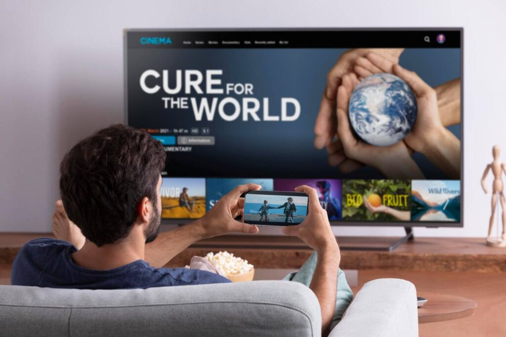 A person relaxes on a couch, watching a documentary on TV while streaming a high quality video on their smartphone, with popcorn nearby.