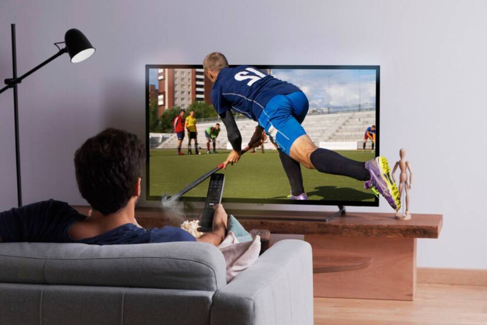 A person relaxes on a couch watching a hockey game on TV, holding a remote.