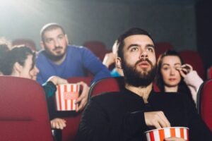 A man enjoying popcorn while watching a movie , seated on red chair with dim lighting.