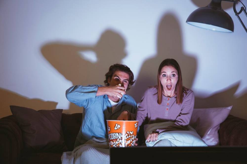 A couple sits on a couch, watching horror movie and enjoying popcorn.