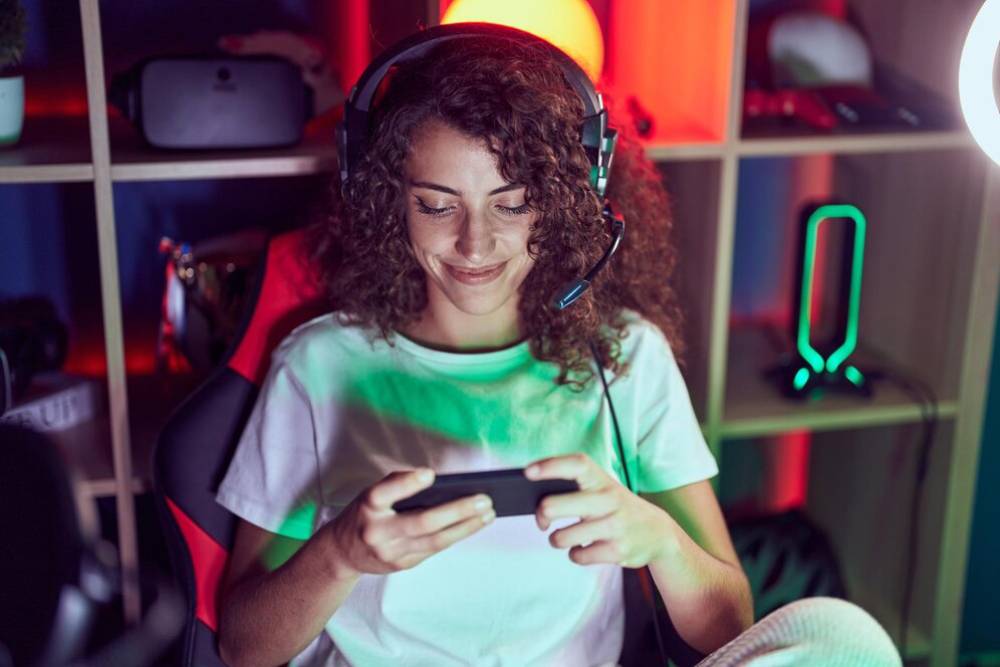 Happy young woman wearing headphones and playing games on her smartphone in a colorful, neon-lit room.